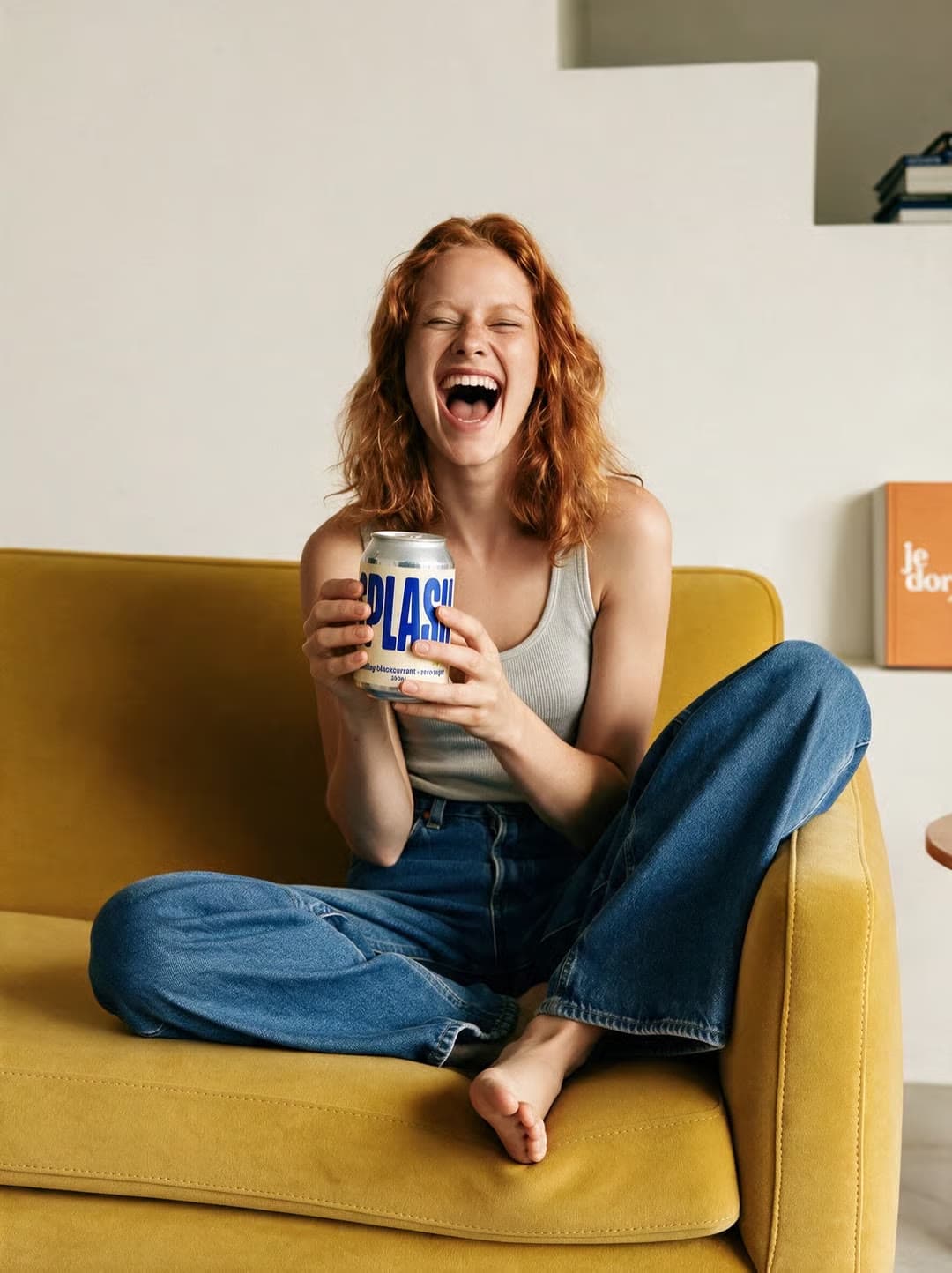 A laughing woman on a yellow sofa holds a drink can