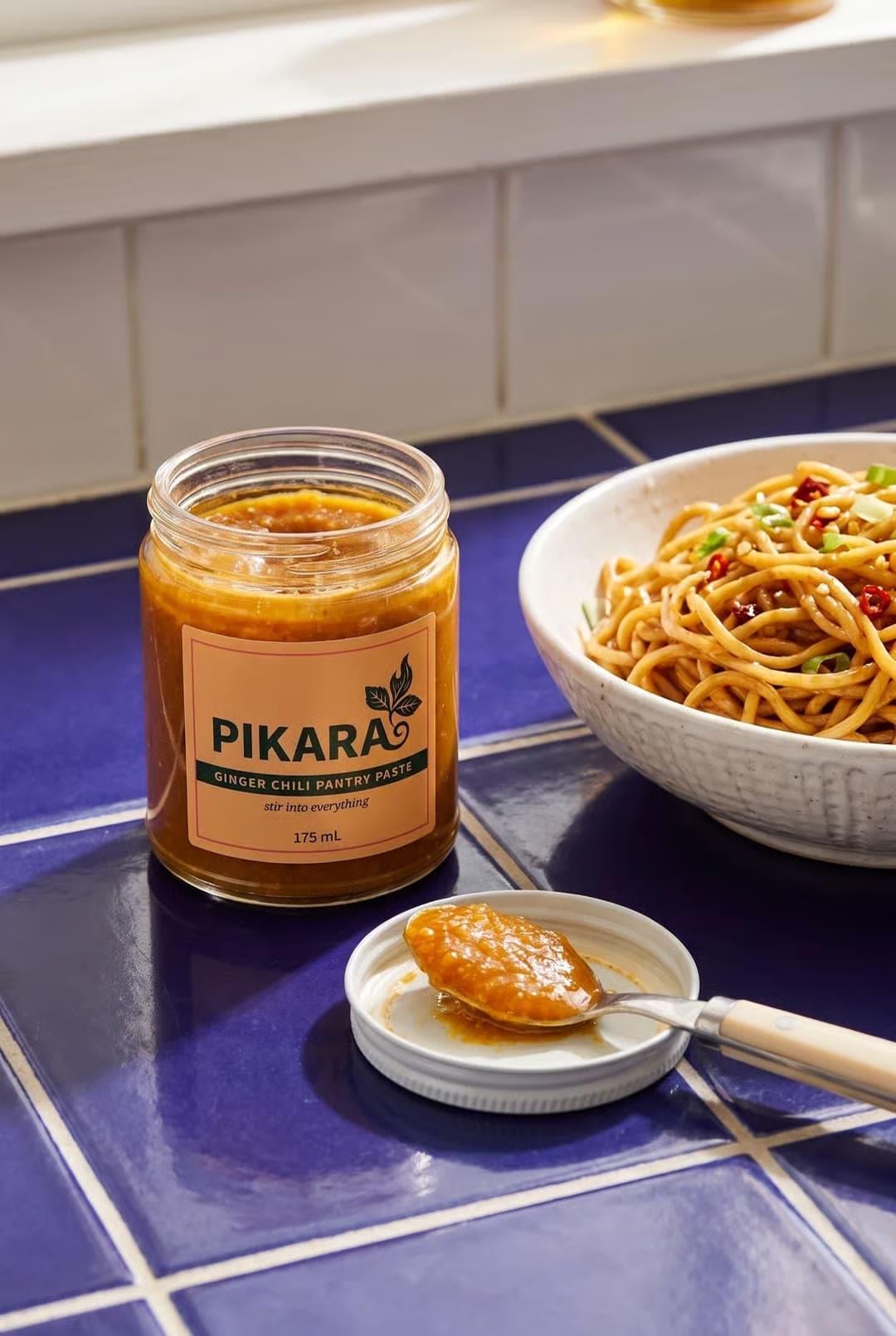 A jar of Pikara chili sauce beside a bowl of noodles on blue tiles
