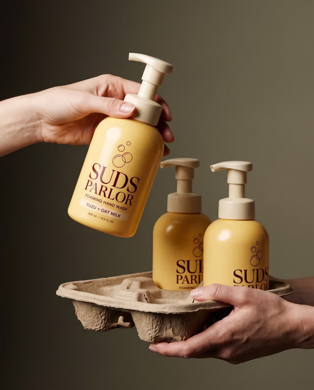 Hands lift three Suds Parlor soap bottles from a cardboard carrier